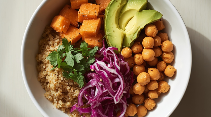 Square overhead shot of a white bowl filled with quinoa, roasted sweet potato, chickpeas, sliced avocado, and shredded red cabbage, garnished with fresh herbs, on a light wooden surface
