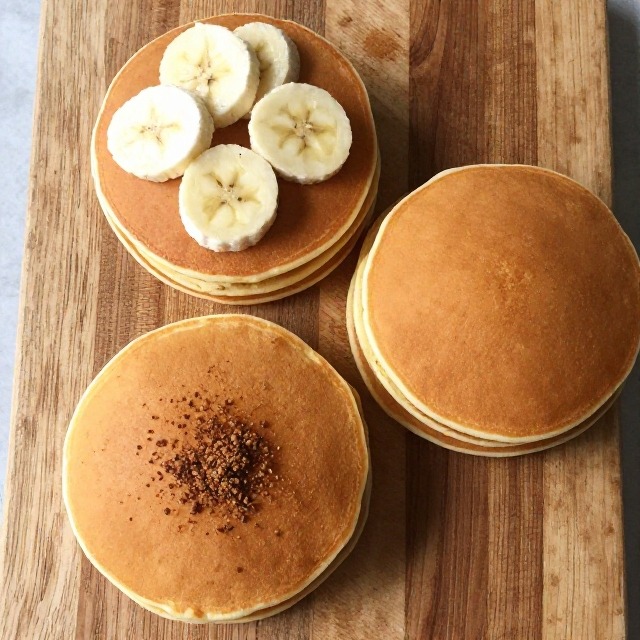 Overhead shot of three cottage cheese pancake variations on a wooden board, one topped with banana slices, one with pumpkin spice dusting, one plain stack, no text, no labels, no packaging, no brand names, no background drinks or glasses of liquid
