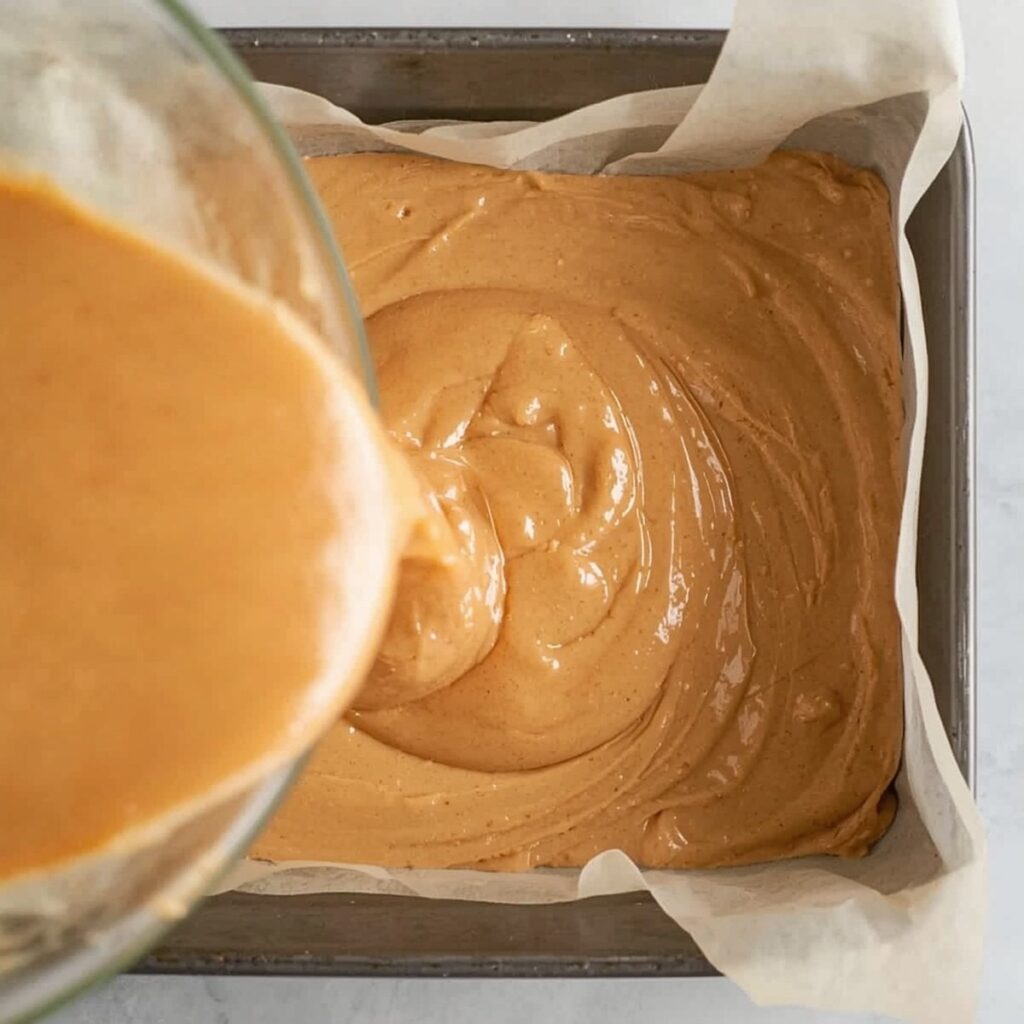 Peanut butter fudge with marshmallow fluff being poured into a parchment-lined baking pan, no text or branding